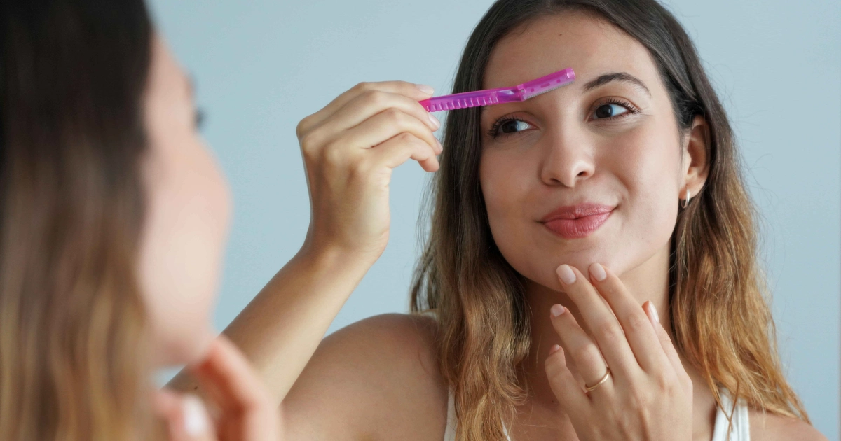 Young woman in Hollidaysburg, PA using a pink eyebrow razor near her eyebrow, reflected in a mirror, likely for dermaplane or shaping.